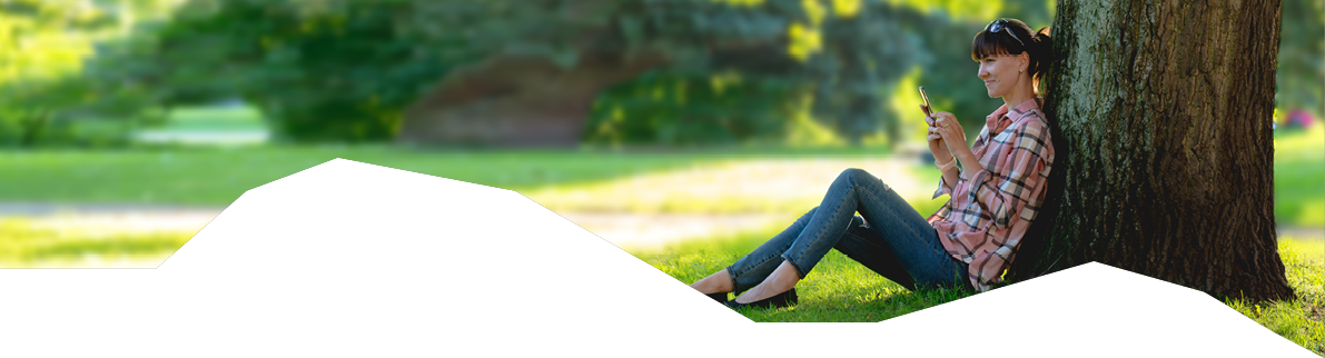 young woman sits under a tree with a phone in hands in the summer park 